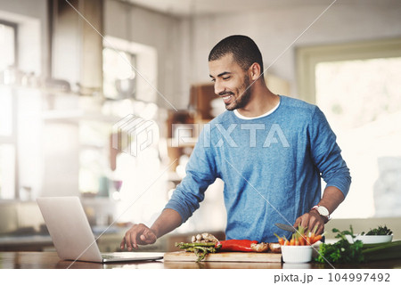 Young casual man in the kitchen on a laptop while preparing a healthy meal at home. Happy smiling male browsing and learning on computer on how to cook. Guy alone checking online recipes on the web. Young casual man in the kitchen on a laptop while preparing a healthy meal at home. Happy smiling male browsing and learning on computer on how to cook. Guy alone checking online recipes on the web. 104997492