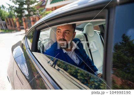 A caucasian man in a blue suit looks out the open window while sitting in the back seat of a car. Business class passenger.  104998190