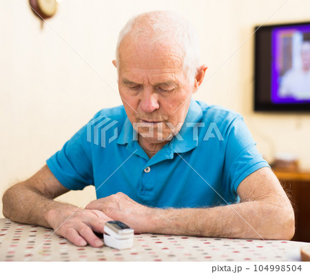 Focused worried elderly man measuring himself oxygen saturation while sitting at home table, using pulse oximeter. Health self-control concept Focused worried elderly man measuring himself oxygen saturation while sitting at home table, using pulse oximeter. Health self-control concept 104998504