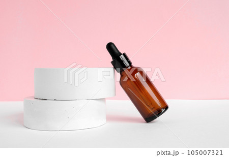 Amber glass dropper cosmetic bottle, white podium. Natural skin care SPA beauty product design. Mineral organic oil on pink background. Mock-Up. Oily pipette. Face and body treatment. Front view 105007321
