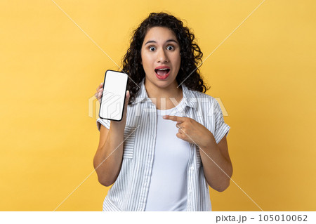 Portrait of amazed surprised woman with dark wavy hair holding pointing ar mobile phone with blank screen, mockup for advertisement. Indoor studio shot isolated on yellow background. 105010062