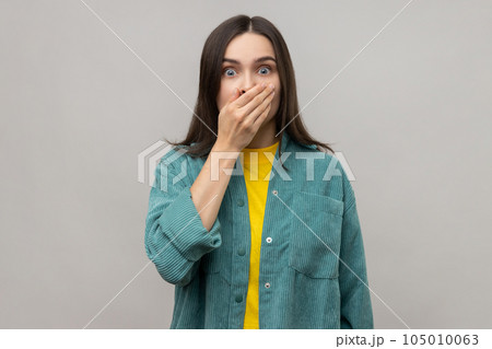 Portrait of scared shocked dark haired woman closing mouth with hand, afraid to say too much, embarrassment, wearing casual style jacket. Indoor studio shot isolated on gray background. 105010063
