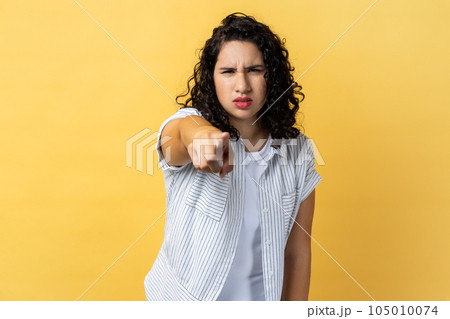 Portrait of strict woman with dark wavy hair noticing and pointing finger to camera, accusing with serious bossy face, making choice. Indoor studio shot isolated on yellow background. 105010074