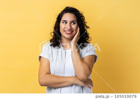 Portrait of dreamy beautiful woman with dark wavy hair standing and dreaming, making wish, love, looking away with toothy smile.Indoor studio shot isolated on yellow background. 105010079