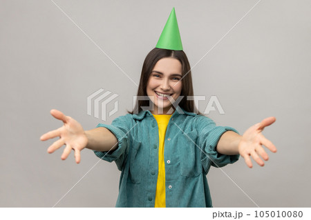 Woman with party cone keeping hands wide open to embrace, giving warm welcome, greeting and sharing love, wearing casual style jacket. Indoor studio shot isolated on gray background. 105010080