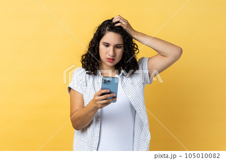Portrait of pensive beautiful young adult woman with dark wavy hair using cell phone thinking what to answer, keeps hand on head. Indoor studio shot isolated on yellow background. 105010082