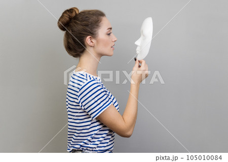 Side view portrait of beautiful dark haired woman with bun hairstyle wearing striped T-shirt standing holding and looking at white mask. Indoor studio shot isolated on gray background. 105010084
