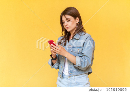 Side view portrait of serious concentrated woman wearing denim jacket using cell phone, chatting with somebody with attentive expression. Indoor studio shot isolated on yellow background. Side view portrait of serious concentrated woman wearing denim jacket using cell phone, chatting with somebody with attentive expression. Indoor studio shot isolated on yellow background. 105010096