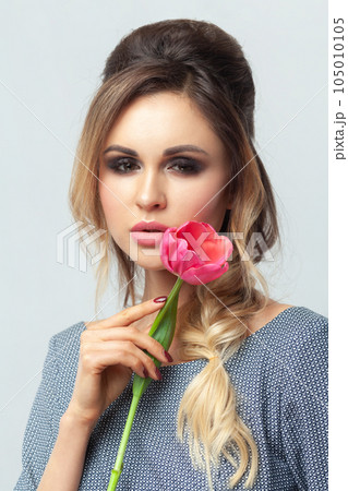 Closeup portrait of attractive gentle blonde woman fashion model with makeup and hairstyle, holding tulip near her face, wearing grey elegant dress. Indoor studio shot isolated on gray background. Closeup portrait of attractive gentle blonde woman fashion model with makeup and hairstyle, holding tulip near her face, wearing grey elegant dress. Indoor studio shot isolated on gray background. 105010105