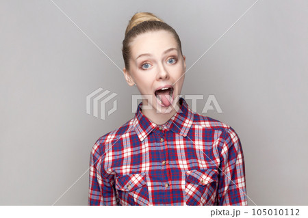 Portrait of foolish crazy playful woman with bun hairstyle looking at camera with open mouth and showing tongue out, wearing checkered shirt. Indoor studio shot isolated on gray background. 105010112