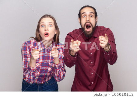 Portrait of shocked amazed woman and man standing together and pointing at camera, choosing you, looking at camera with big eyes. Indoor studio shot isolated on gray background. Portrait of shocked amazed woman and man standing together and pointing at camera, choosing you, looking at camera with big eyes. Indoor studio shot isolated on gray background. 105010115