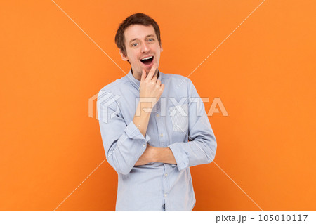 Portrait of excited amazed young adult man standing with hand on chin, having good idea, looking at camera, wearing light blue shirt. Indoor studio shot isolated on orange background. Portrait of excited amazed young adult man standing with hand on chin, having good idea, looking at camera, wearing light blue shirt. Indoor studio shot isolated on orange background. 105010117