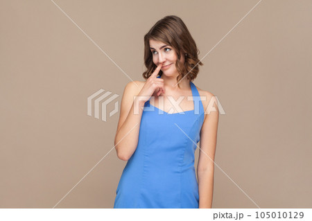 Woman with wavy hair drilling nose fooling around, showing tongue out and picking nose with comical silly expression, wearing blue dress. Indoor studio shot isolated on light brown background. 105010129
