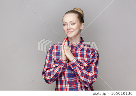 Portrait of sly attractive woman with bun hairstyle keeps palms together, planning devil plan or prank, wearing checkered shirt. Indoor studio shot isolated on gray background. Portrait of sly attractive woman with bun hairstyle keeps palms together, planning devil plan or prank, wearing checkered shirt. Indoor studio shot isolated on gray background. 105010130