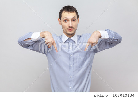 Portrait of serious strict young adult man standing pointing both index fingers down, saying here and right now, wearing light blue shirt. Indoor studio shot isolated on gray background. Portrait of serious strict young adult man standing pointing both index fingers down, saying here and right now, wearing light blue shirt. Indoor studio shot isolated on gray background. 105010134