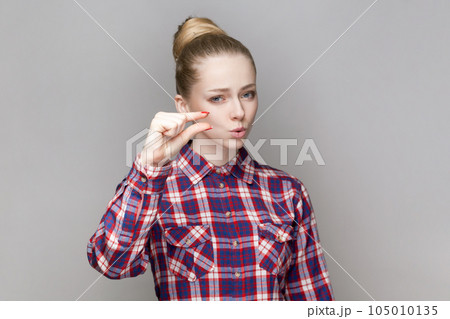 Portrait of beautiful adorable woman with bun hairstyle standing looking at camera, showing small gesture with fingers, wearing checkered shirt. Indoor studio shot isolated on gray background. Portrait of beautiful adorable woman with bun hairstyle standing looking at camera, showing small gesture with fingers, wearing checkered shirt. Indoor studio shot isolated on gray background. 105010135