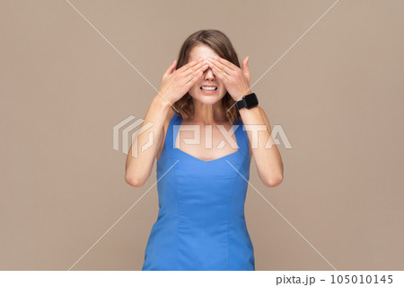 Don't want to look at this. Portrait of scared woman with wavy hair standing, covering eyes with hand, refusing to watch, wearing blue dress. Indoor studio shot isolated on light brown background. Don't want to look at this. Portrait of scared woman with wavy hair standing, covering eyes with hand, refusing to watch, wearing blue dress. Indoor studio shot isolated on light brown background. 105010145