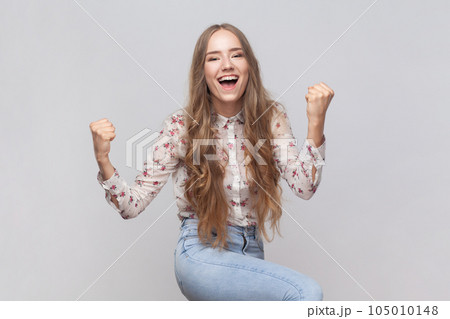 I am champion. Portrait of woman with wavy blond hair screaming for joy and raising high her fists, celebrating successful winning, incredible victory. Indoor studio shot isolated on gray background. I am champion. Portrait of woman with wavy blond hair screaming for joy and raising high her fists, celebrating successful winning, incredible victory. Indoor studio shot isolated on gray background. 105010148