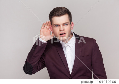Young man holding hand near ear trying to hear secrets and find out gossips, listen to private whispers, , wearing violet suit and white shirt. Indoor studio shot isolated on grey background. 105010166