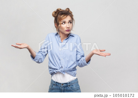 Who knows. Clueless hesitant beautiful blonde woman spreads palms sideways shrugs shoulders has indecisive expression, looking away, wearing blue shirt. Indoor studio shot isolated on gray background. Who knows. Clueless hesitant beautiful blonde woman spreads palms sideways shrugs shoulders has indecisive expression, looking away, wearing blue shirt. Indoor studio shot isolated on gray background. 105010172