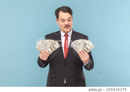 Portrait of rich man with mustache standing counting money., holding big fans of dollar banknotes, wearing black suit with red tie. Indoor studio shot isolated on light blue background. 105010175