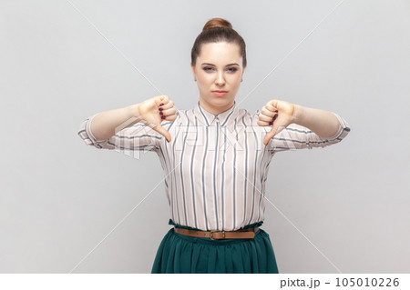 Portrait of dissatisfied displeased attractive woman wearing striped shirt and green skirt standing showing dislike gesture, looks unhappy. Indoor studio shot isolated on gray background. Portrait of dissatisfied displeased attractive woman wearing striped shirt and green skirt standing showing dislike gesture, looks unhappy. Indoor studio shot isolated on gray background. 105010226