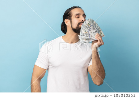 Portrait of rich man with beard wearing white T-shirt holding big fan of money, smelling dollars banknotes with pleasure. Indoor studio shot isolated on blue background. Portrait of rich man with beard wearing white T-shirt holding big fan of money, smelling dollars banknotes with pleasure. Indoor studio shot isolated on blue background. 105010253
