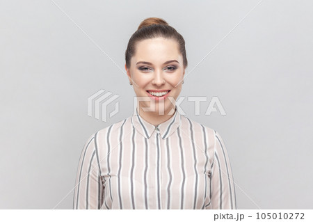 Portrait of attractive joyful cheerful woman wearing striped shirt looking at camera with toothy smile, being in good mood, expressing positiveness. Indoor studio shot isolated on gray background. Portrait of attractive joyful cheerful woman wearing striped shirt looking at camera with toothy smile, being in good mood, expressing positiveness. Indoor studio shot isolated on gray background. 105010272
