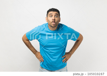 Portrait of funny crazy childish unshaven man wearing blue T- shirt standing with hands on hips, showing tongue out, demonstrates playful behavior. Indoor studio shot isolated on gray background. 105010287