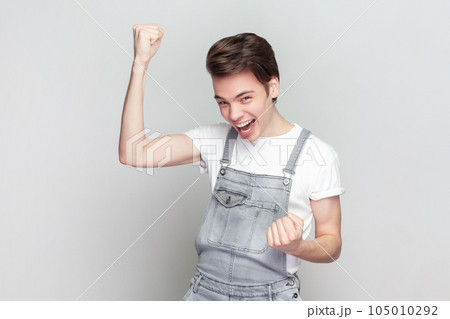 Yes, finally success. Brunette man raises arms with clenched fists, celebrates something has upbeat mood, feels euphoric, wearing denim overalls. Indoor studio shot isolated on gray background. 105010292