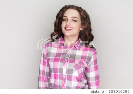 Portrait of woman with curly hair being very glad smiling with broad smile showing her perfect teeth, having fun, wearing pink checkered shirt. Indoor studio shot isolated on gray background. 105010293