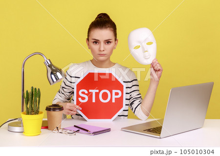 Portrait of serious dark haired woman holding white mask with unknown face and red traffic sign while sitting on workplace with laptop. Indoor studio studio shot isolated on yellow background. Portrait of serious dark haired woman holding white mask with unknown face and red traffic sign while sitting on workplace with laptop. Indoor studio studio shot isolated on yellow background. 105010304