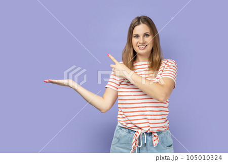 Portrait of blond woman wearing striped T-shirt presenting advertising area on her palm and pointing to copy space, holding empty place for commercial. Indoor studio shot isolated on purple background Portrait of blond woman wearing striped T-shirt presenting advertising area on her palm and pointing to copy space, holding empty place for commercial. Indoor studio shot isolated on purple background 105010324
