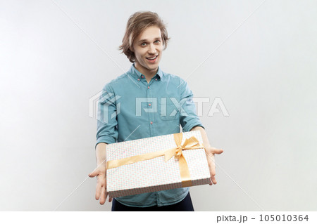 Portrait of satisfied friendly handsome young man standing with gift in hands, giving present to her girl friend on birthday, wearing blue shirt. Indoor studio shot isolated on gray background. Portrait of satisfied friendly handsome young man standing with gift in hands, giving present to her girl friend on birthday, wearing blue shirt. Indoor studio shot isolated on gray background. 105010364
