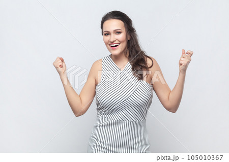 Emotional brunette woman raises clenched fists, exclaims with excitement, rejoices sweet success, feels taste of victory, wearing striped dress. Indoor studio shot isolated on gray background. 105010367