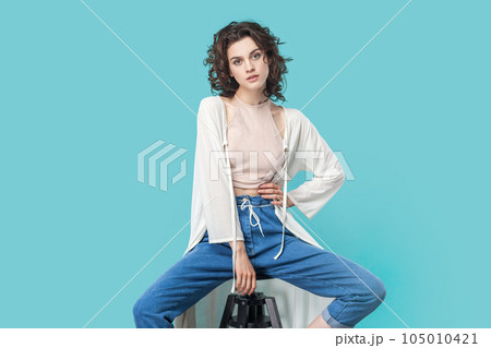 Portrait of winsome young adult woman with wavy hair sitting on chair, looking at camera confident expression, wearing stylish outfit. Indoor studio shot isolated on blue background. Portrait of winsome young adult woman with wavy hair sitting on chair, looking at camera confident expression, wearing stylish outfit. Indoor studio shot isolated on blue background. 105010421
