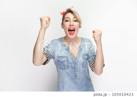 Portrait of extremely happy cheerful blonde woman wearing blue denim shirt and red headband standing clenched fists, celebrating victory. Indoor studio shot isolated on gray background. Portrait of extremely happy cheerful blonde woman wearing blue denim shirt and red headband standing clenched fists, celebrating victory. Indoor studio shot isolated on gray background. 105010423