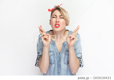 Portrait of hopeful attractive blonde woman wearing blue denim shirt and red headband standing with crossed fingers, praying for good luck. Indoor studio shot isolated on gray background. 105010431