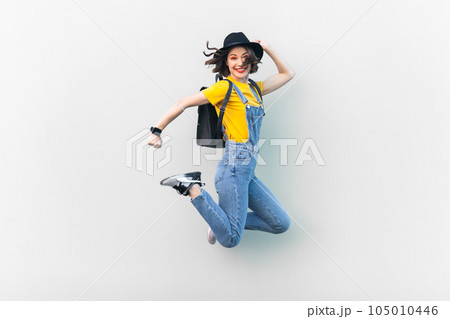 Portrait of attractive overjoyed hipster woman in blue denim overalls, yellow T-shirt and black hat, jumping, being in good mood, expressing happiness. Indoor studio shot isolated on gray background. 105010446