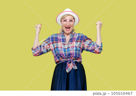 Overjoyed senior woman in checkered shirt, hat, eyeglasses clenches fists with happiness, opens mouth widely as shouts loudly, celebrates her success. Indoor studio shot isolated on yellow background. Overjoyed senior woman in checkered shirt, hat, eyeglasses clenches fists with happiness, opens mouth widely as shouts loudly, celebrates her success. Indoor studio shot isolated on yellow background. 105010467