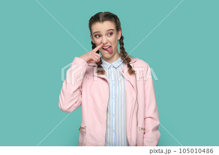 Portrait of funny childish teenager girl with braids wearing pink jacket standing with finger in nose, sticking tongue out, looking away. Indoor studio shot isolated on green background. 105010486