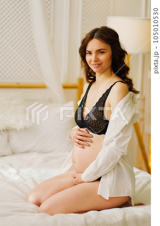 a slender pregnant woman in a black underwea and white shirt on the bed 105010530
