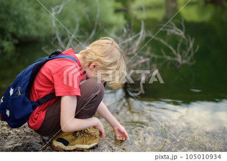 Preteen boy in red shirt is exploring nature and playing with water in lake during hiking in mountains valley. Active leisure for energetic children. Summer outdoor recreation for kids 105010934
