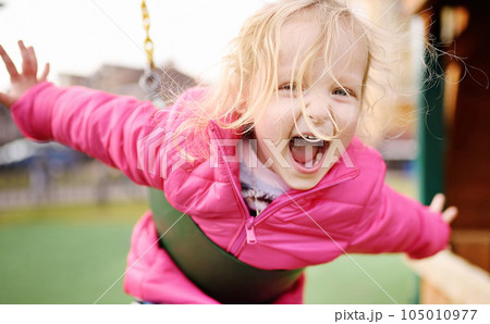 Cute little girl having fun on outdoor playground. Spring or summer active sport leisure for kids. Child on plastic swing 105010977