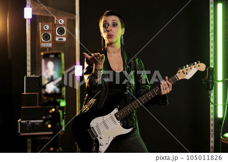 Rockstar blowing air kisses in front of smartphone camera while playing at electric guitar in sound studio, working at rock album using electricinstrument. Rebel musician recording heavy metal song 105011826