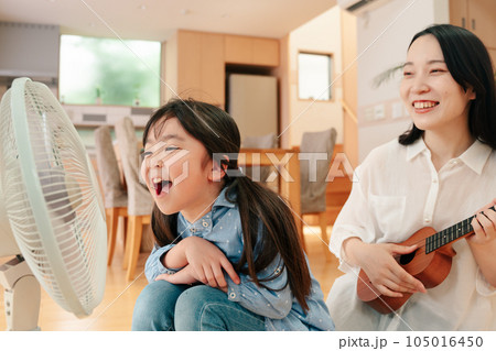 扇風機に向かって大声で歌う女の子とウクレレを弾く母親 105016450