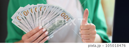 Manager woman holding stack of dollar bills and showing thumb up gesture while sitting at office desk. 105018476