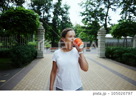 Smiling woman tourist walking in the park alley on a summer day, holding orange cardboard mug with takeaway hot drink 105020797