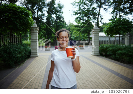 Smiling beautiful confident woman tourist walking in the park alley, holding orange cardboard mug with takeaway drink Smiling beautiful confident woman tourist walking in the park alley, holding orange cardboard mug with takeaway drink 105020798