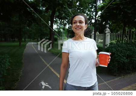 Latin American young smiling pretty woman holding takeaway coffee in paper cup, walking along the alley of a city park Latin American young smiling pretty woman holding takeaway coffee in paper cup, walking along the alley of a city park 105020800